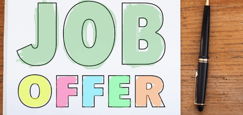 Schriftzug 'Job Offer' in großen bunten Buchstaben auf einem Blatt Papier auf Holzuntergrund, daneben ein Smartphone und ein schwarzer Kugelschreiber