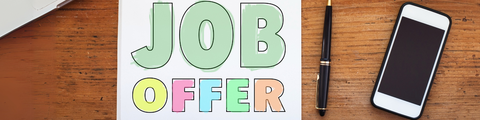 Schriftzug 'Job Offer' in großen bunten Buchstaben auf einem Blatt Papier auf Holzuntergrund, daneben ein Smartphone und ein schwarzer Kugelschreiber