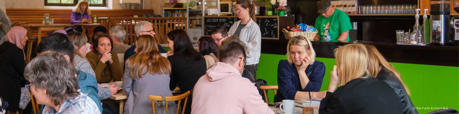 Personen des Deutsch-Cafés im Café Tür an Tür: Menschen mit und ohne Migrationsgeschichte sitzen gemeinsam am Tisch und lernen Deutsch in entspannter Atmosphäre. Bildnachweis: Ran Fuchs https://www.ranfuchs.art Ursula Bué https://www.aerolibrium.de
