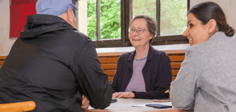 Teilnehmende im Deutsch-Café von Tür an Tür sitzen gemeinsam mit einer Freiwilligen am Tisch, sprechen miteinander und lernen in einer offenen, freundlichen Atmosphäre. Foto: Ran Fuchs und Ursula Boé