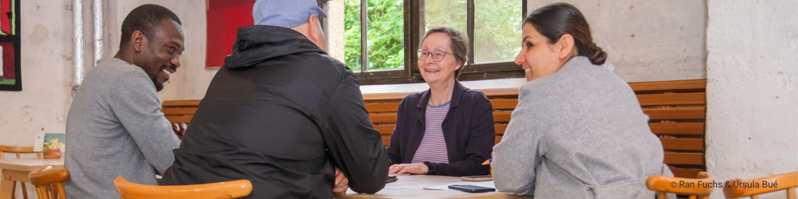Teilnehmende im Deutsch-Café von Tür an Tür sitzen gemeinsam mit einer Freiwilligen am Tisch, sprechen miteinander und lernen in einer offenen, freundlichen Atmosphäre. Foto: Ran Fuchs und Ursula Boé