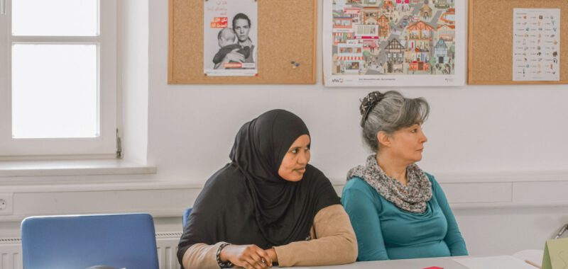 Zwei Teilnehmerinnen sitzen mit Heften im ehrenamtlichen Deutschkurs von Tür an Tür in Augsburg, an der Wand hängen Lernplakate.